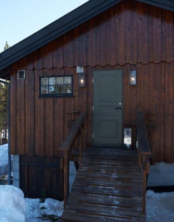 Brun trehytte med trapp opp til grønn ytterdør.