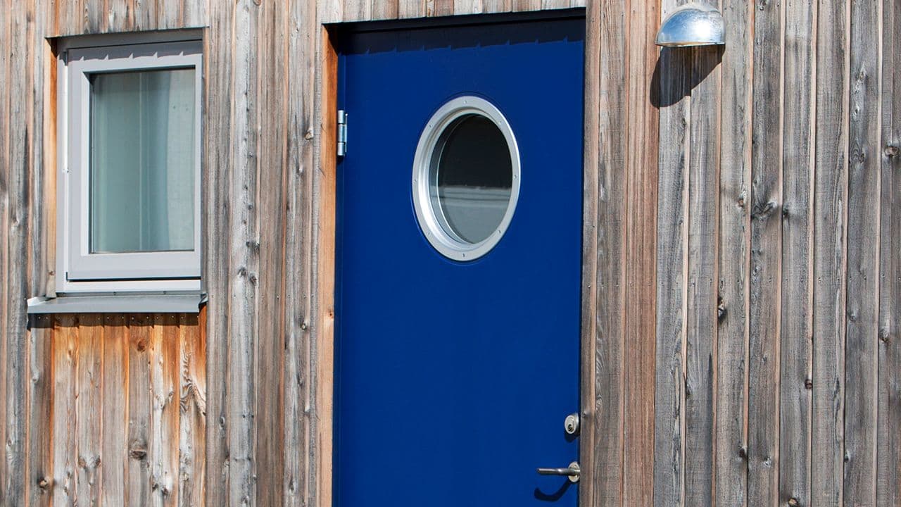 Blå ytterdør med rundt glassfelt og hus med trekledning.